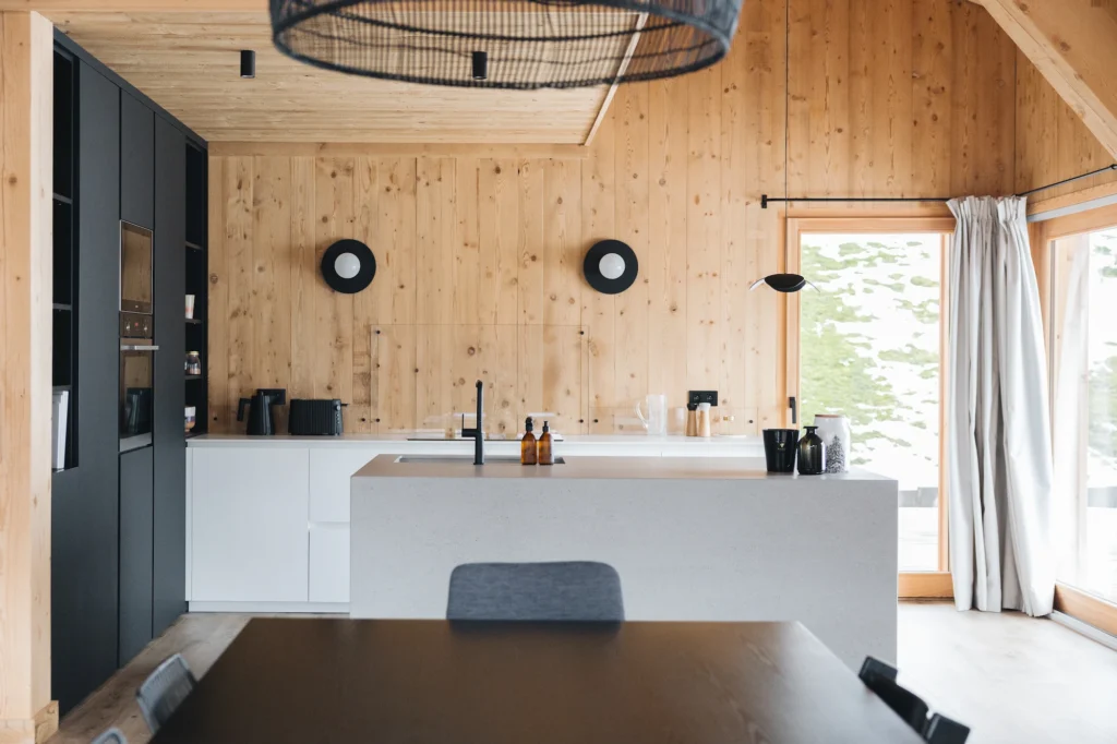 Vue élargie de la cuisine dans le projet Squaw Valley réalisé par InSide Interior Design, agence d’architecture en Haute Savoie. La bouilloire plissée noire est s’intègre parfaitement à la cuisine blanches et aux murs en bois clair.
