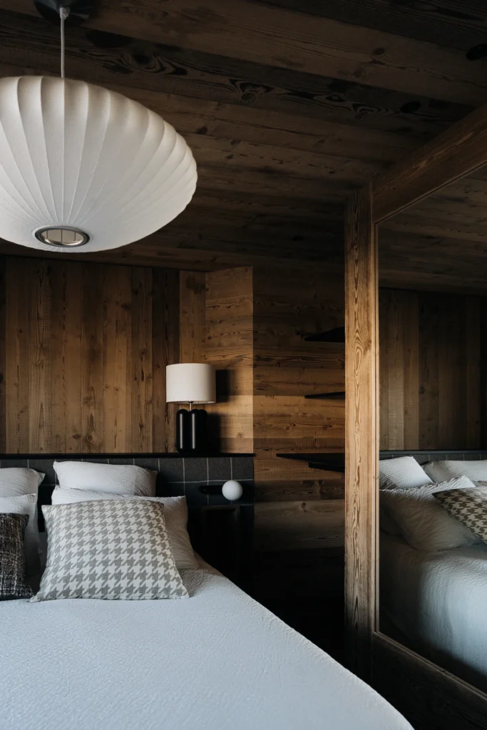 Lampe Charlotte de Cosin vue de loin posée sur une tête de lit feutrée dans une chambre ambiance montagne remplie de bois chaud.