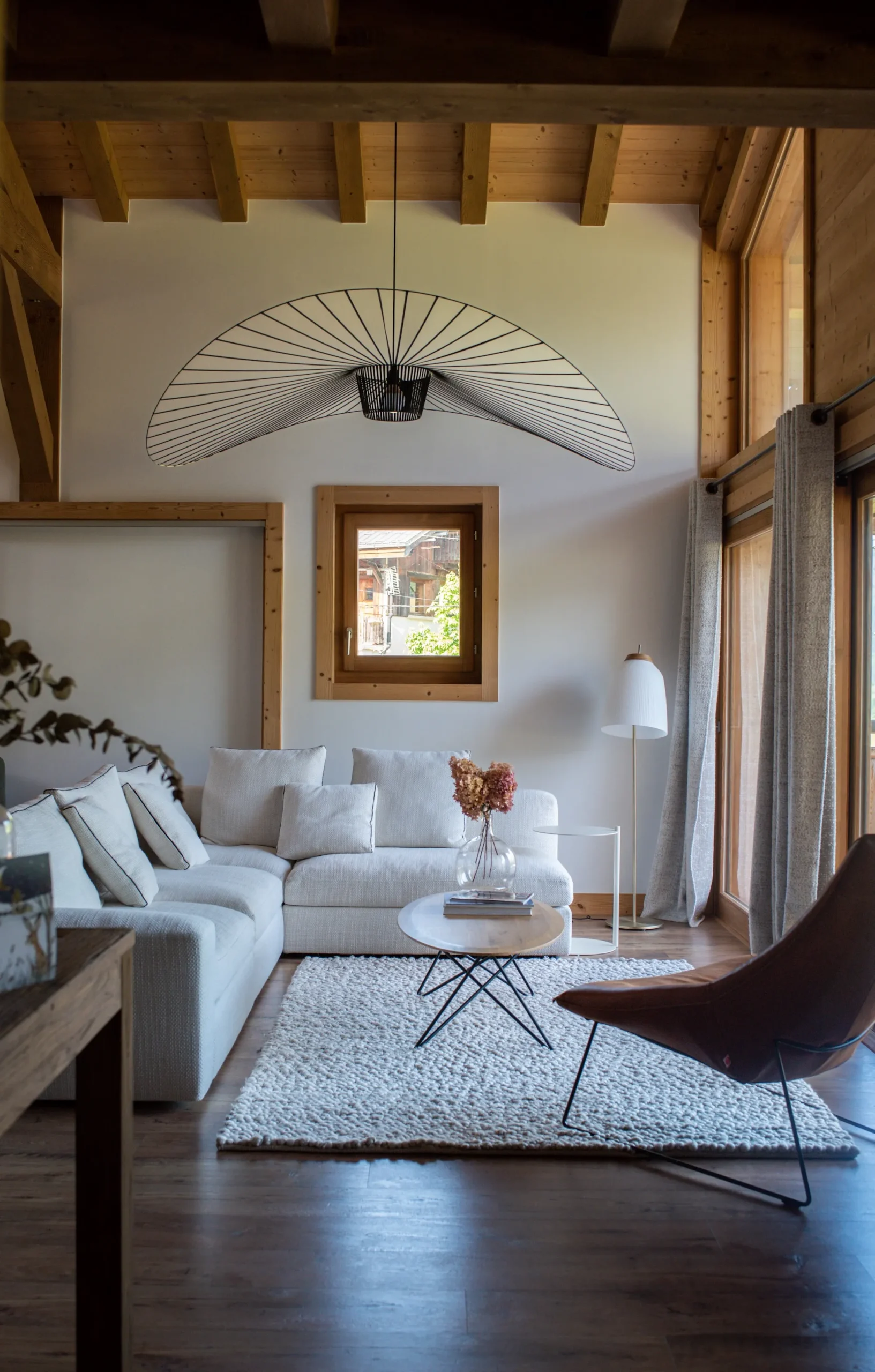 Photo d’un salon aux tons clairs avec un plancher bois de couleur sombre. La suspension est imposante et ancre le salon pour lui donner un style moderne et intemporel. Architecte d’intérieur en Haute-Savoie spécialisée dans les chalets de montage.