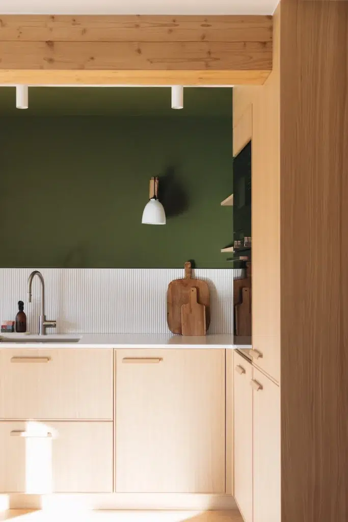 Un mur d’une cuisine est peint en vert, couleur tendance de 2026, pour mettre en avant le bois clair. 