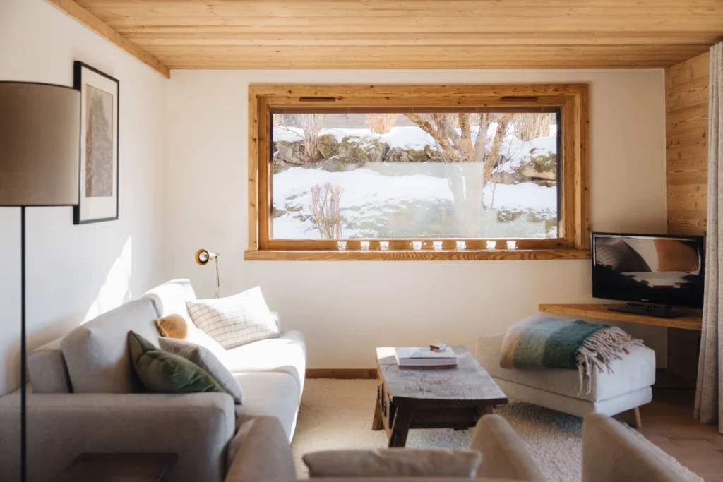 Vue sur le nouveau salon rénové avec une grande fenêtre donnant sur l’extérieur et le paysage enneigé. Le salon est décoré dans un style montagne chic